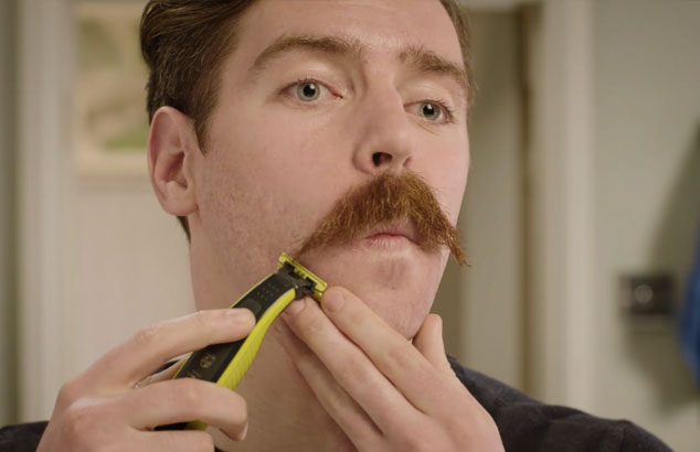 Gesicht eines Mannes mit einem Schnurrbart, der Barthaare auf der Wange mit einem elektrischen Rasierer trimmt.