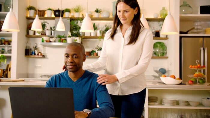 Foto von einem sitzenden Mann, neben ihm steht eine Frau in der Küche und beide schauen auf einen Laptop