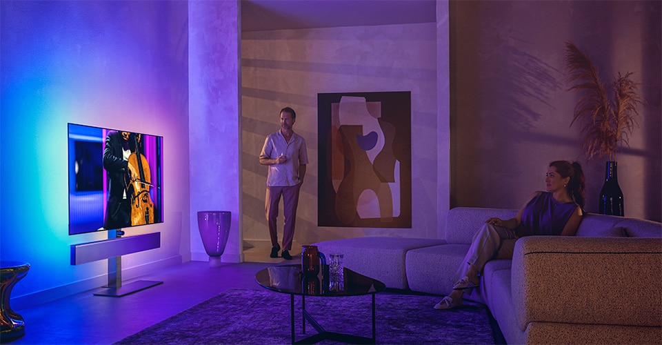 Un homme et une femme dans un salon élégant regardent un téléviseur Philips Ambilight diffusant une performance de violoncelle
