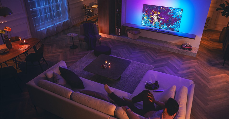 Un homme se détend sur un canapé dans un salon faiblement éclairé, regardant un téléviseur Philips Ambilight