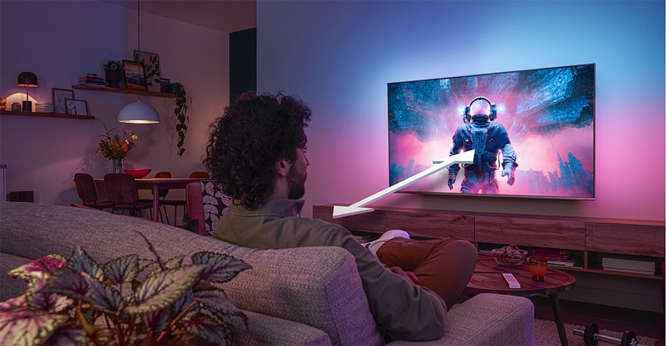 Un homme assis sur un canapé regarde un téléviseur Philips Ambilight affichant une scène d'astronaute colorée, avec une flèche indiquant la distance de visionnage optimale