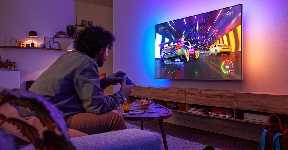 Une personne est assise sur un canapé et joue à un jeu vidéo de course sur un téléviseur Philips Ambilight