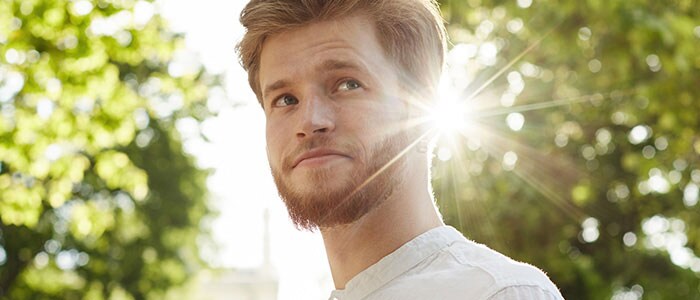 Mann mit kurzem Vollbart und grünen Baumkronen und Sonne im Hintergrund.