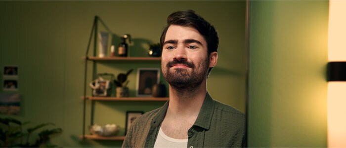 Ein Mann mit dunklem Haar und natürlichem Vollbart sieht lächelnd in den Spiegel in einem Raum mit grünem Hintergrund.